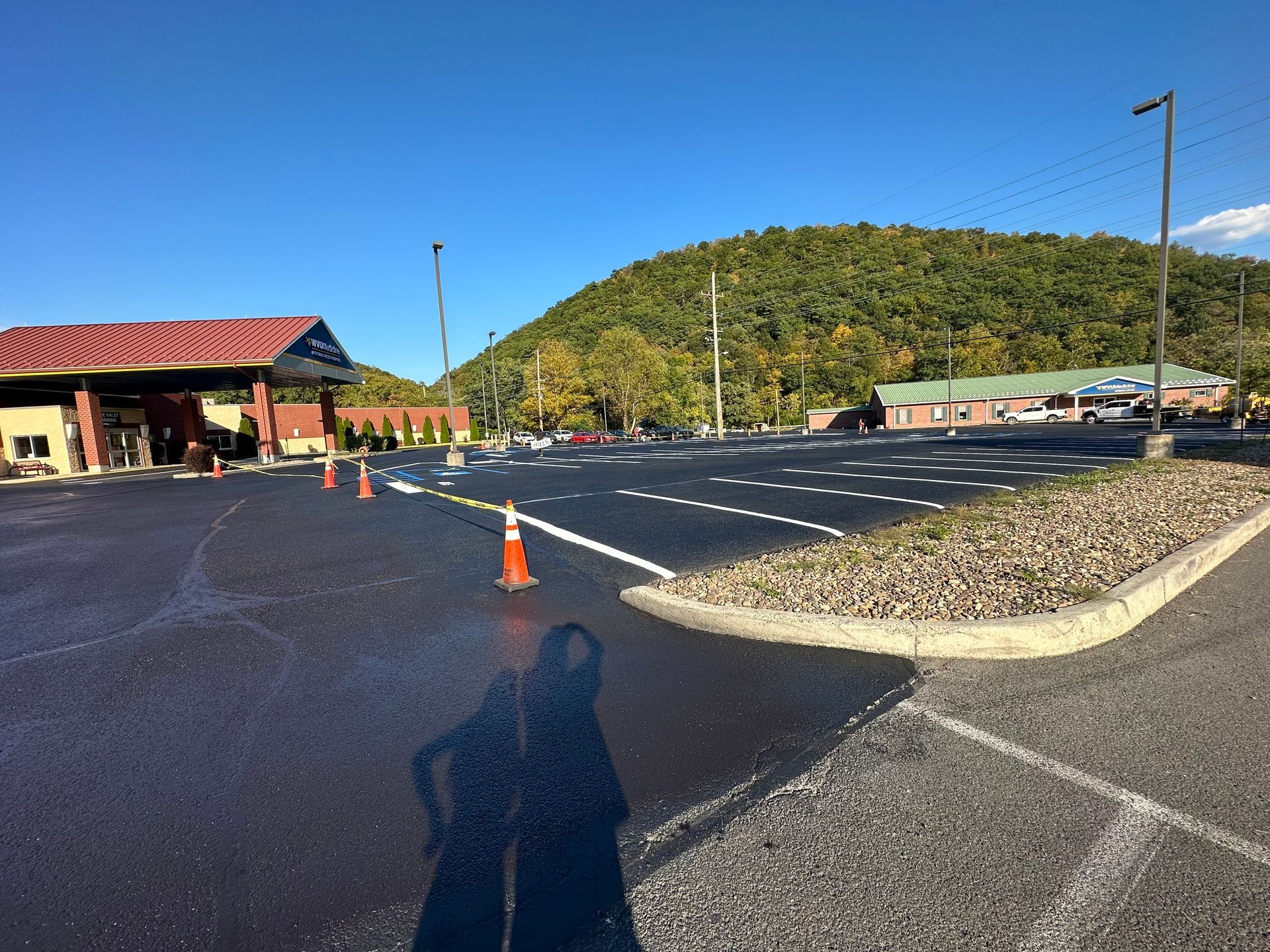 Freshly paved asphalt parking lot with white lines, cones, and buildings, mountain backdrop.