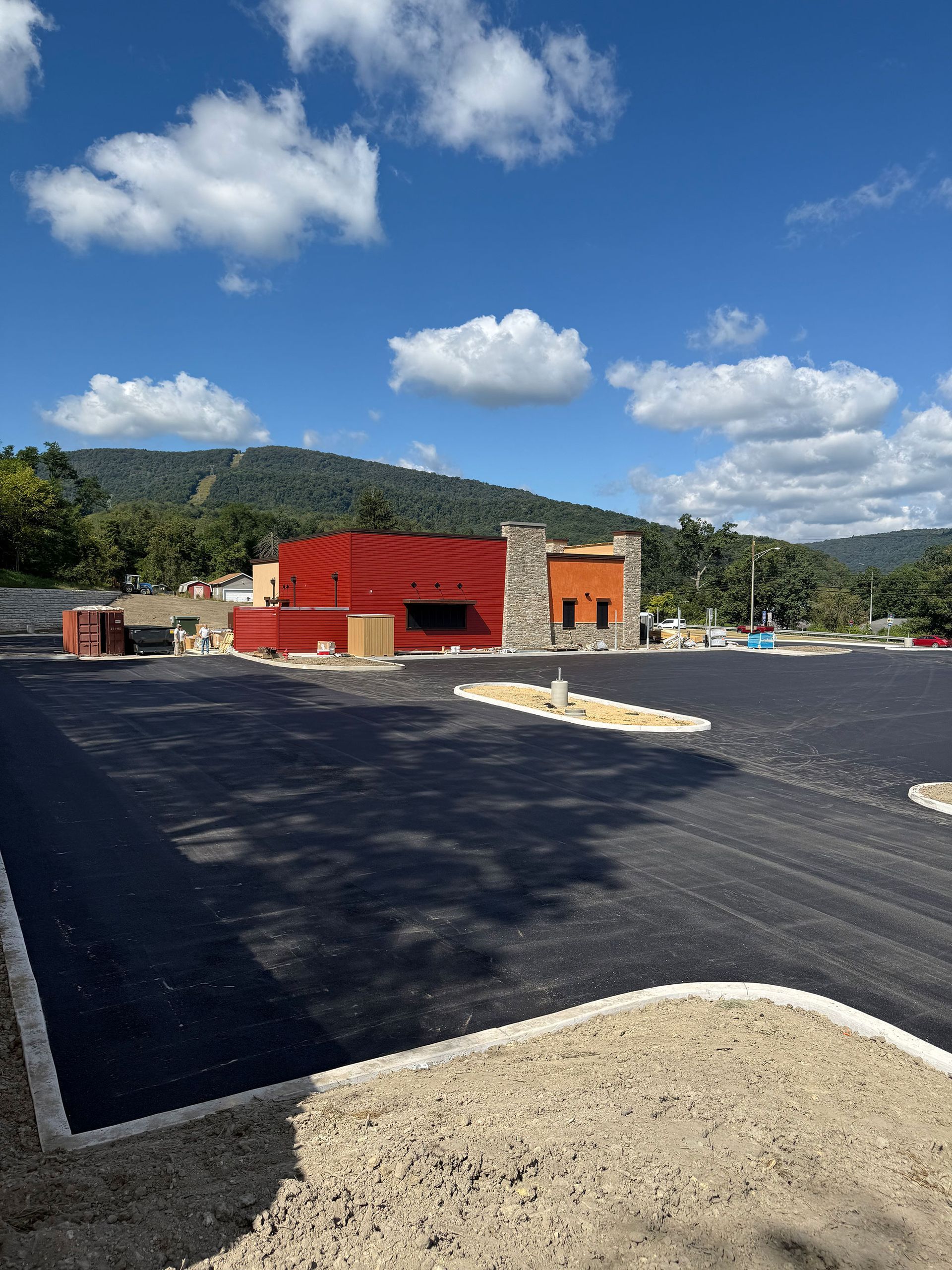 Building under construction with a large paved parking lot, mountains, and blue sky with clouds.