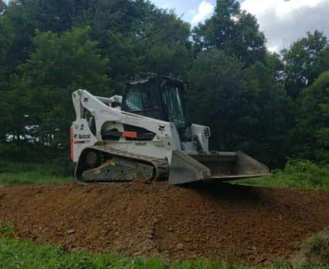 Bobcat skid-steer loader on a dirt pile, in a grassy area with trees in the background.