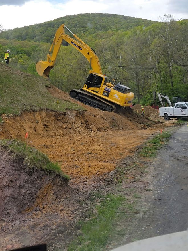 Yellow excavator on a hillside, digging into the earth next to a road, with a white truck.