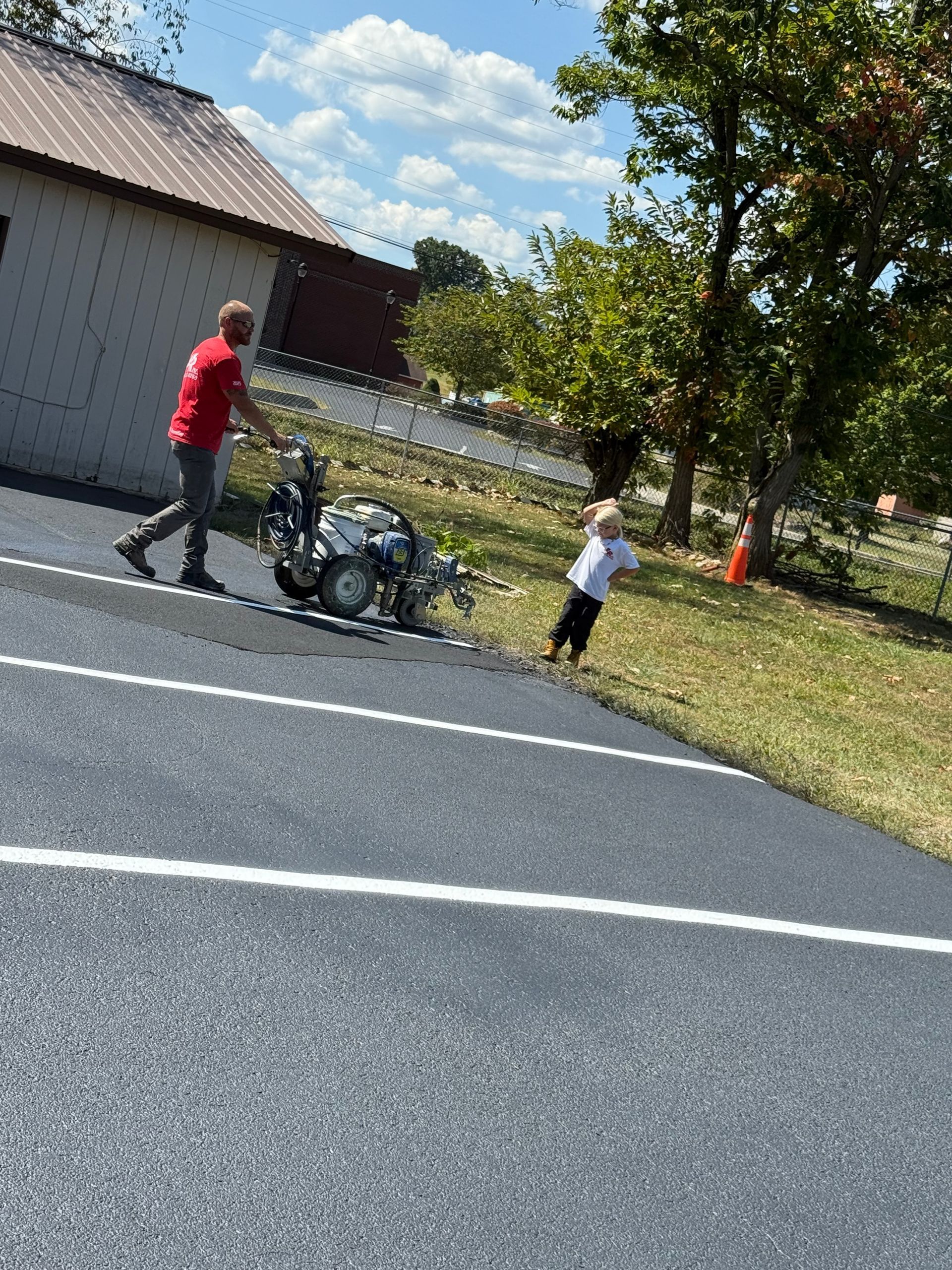 Man in red shirt operating a line painting machine; child walking nearby. Asphalt, grass, building, sunny day.