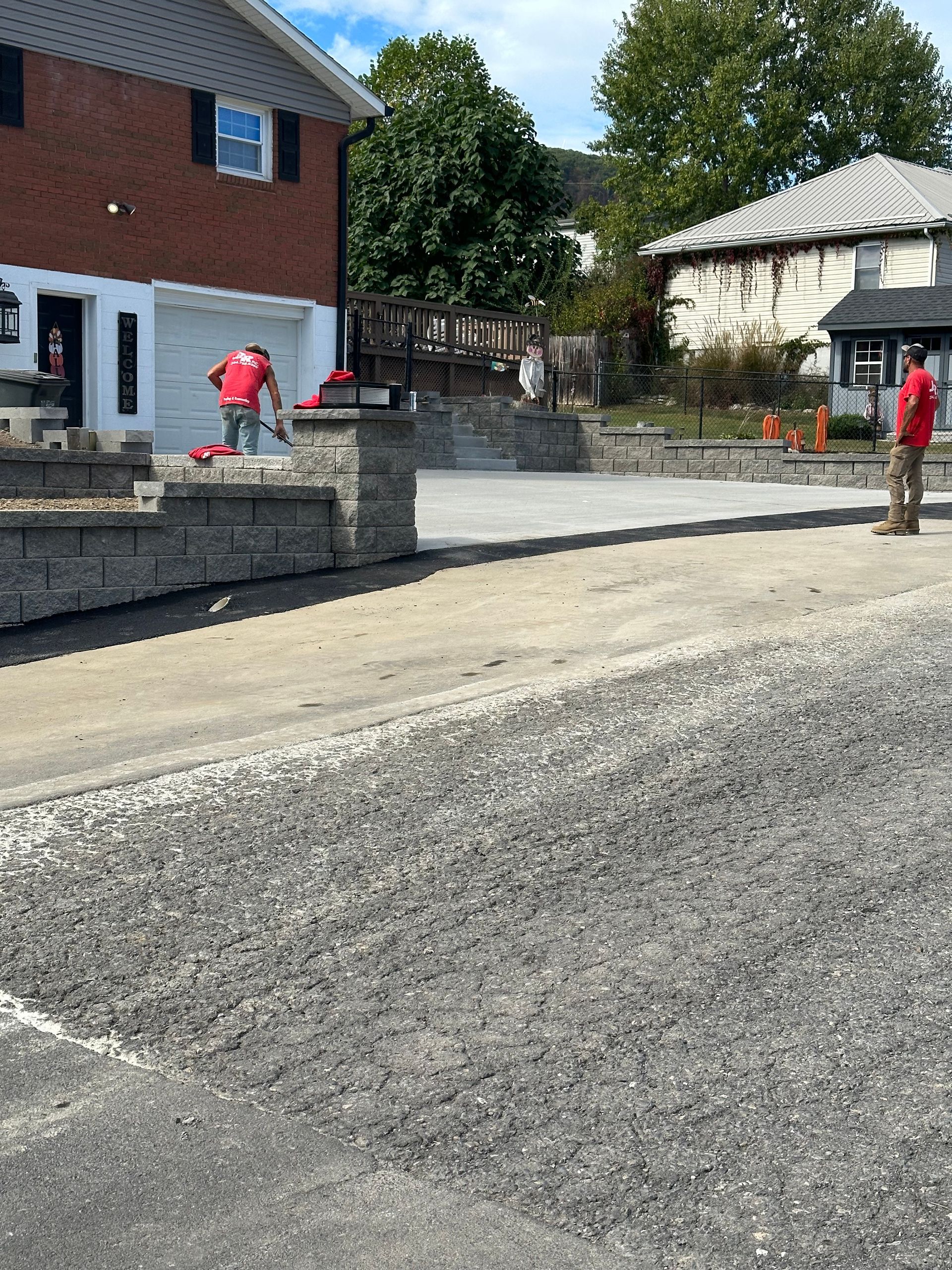 Construction workers paving a driveway with an asphalt roller near a brick building.