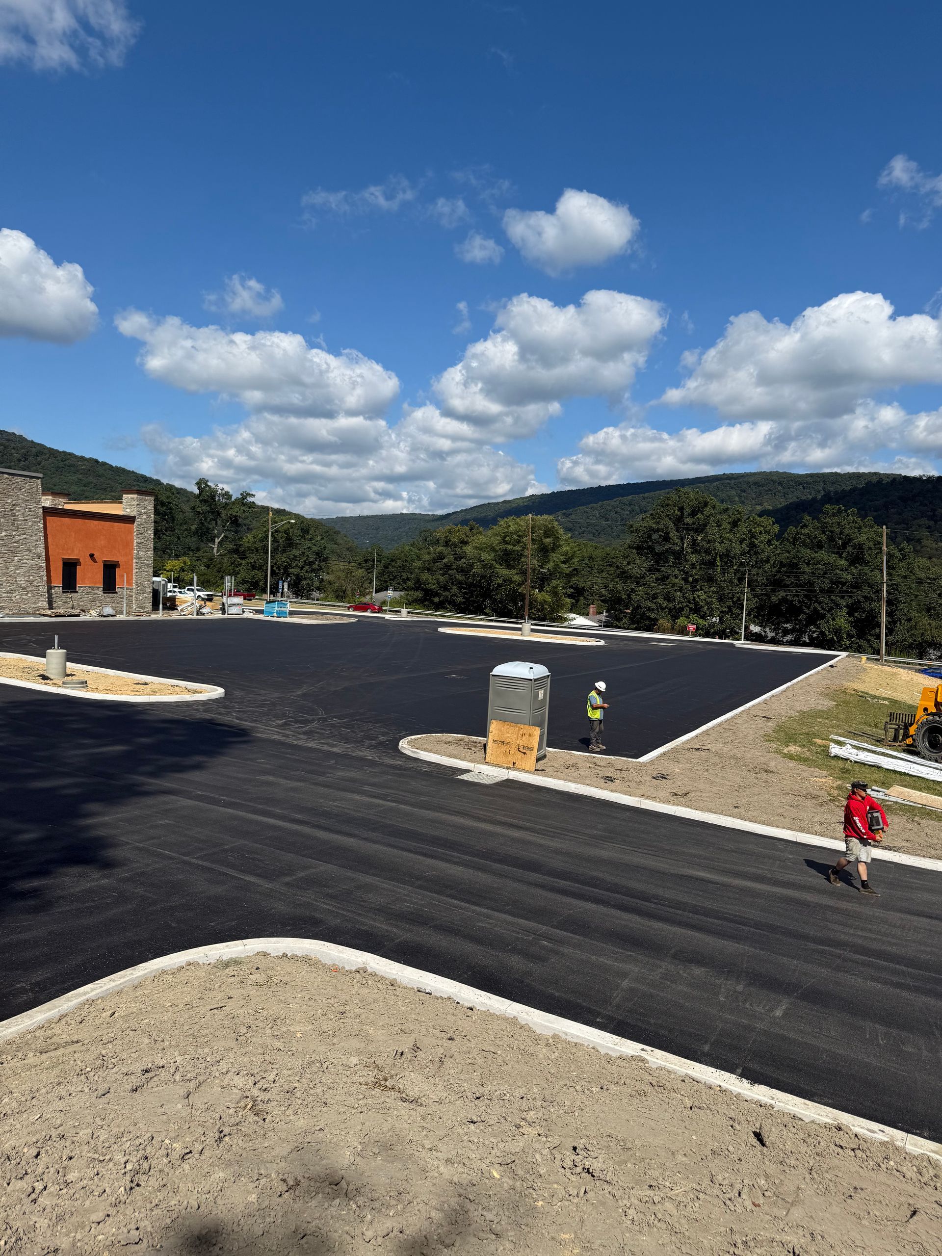 Newly paved parking lot with mountains in background; construction workers and portable toilet visible.