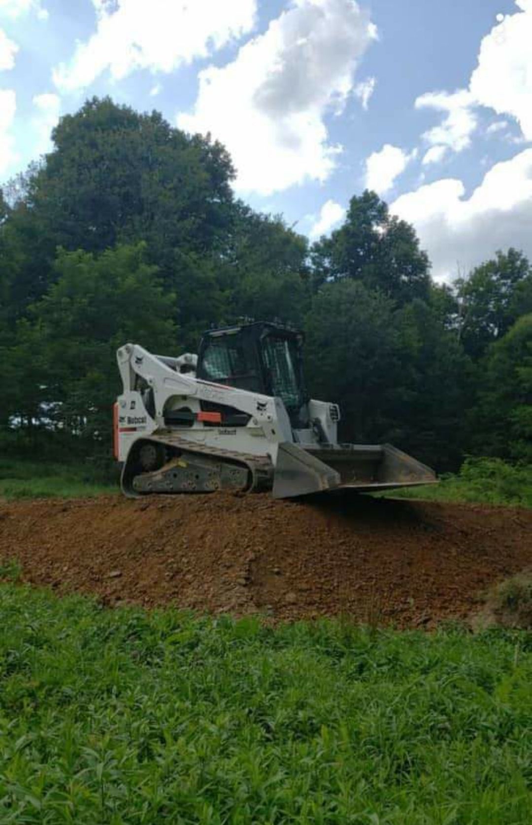 Bobcat construction vehicle on a mound of dirt in a field, trees in the background, blue sky.