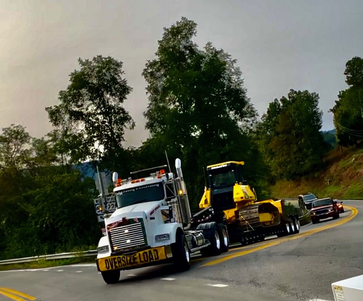 White semi-truck transporting yellow bulldozer on a winding road with overcast sky. 