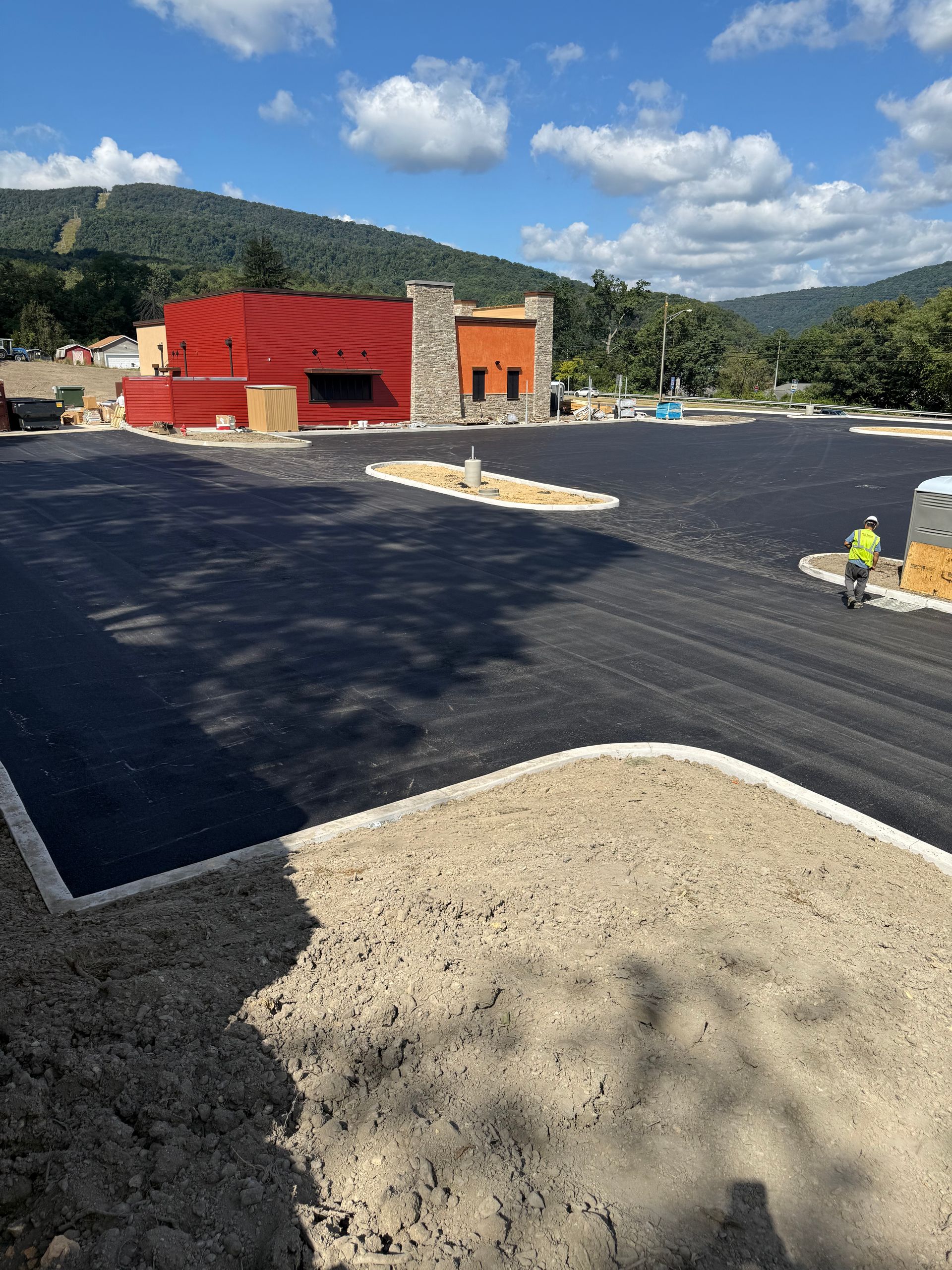 Newly paved parking lot with a red and tan building in the background; a worker stands near a construction vehicle.