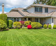 Two-story house with green lawn, trimmed bushes, and colorful flowers under a cloudy sky.
