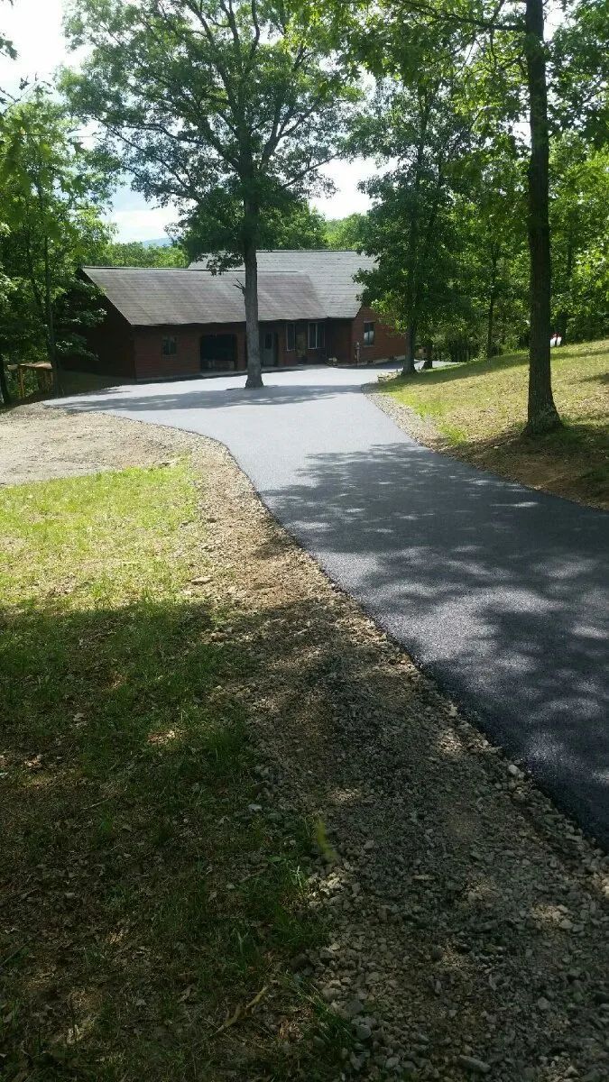 Asphalt driveway leads to a wooden house surrounded by trees.