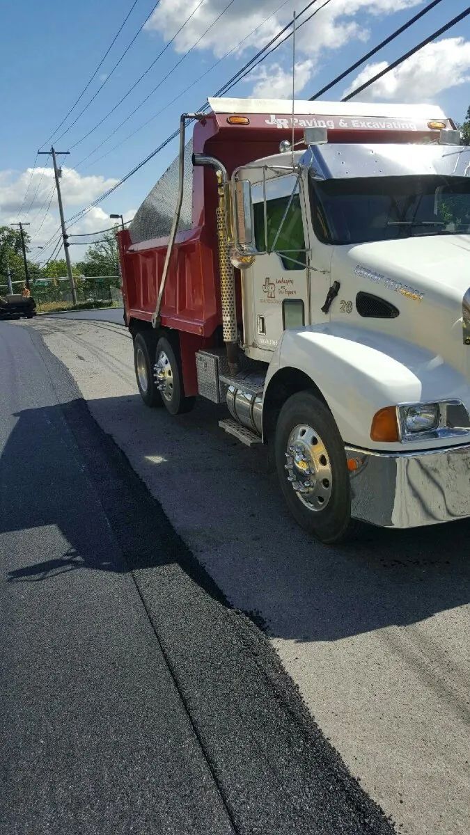 Dump truck on asphalt road during paving. Red truck bed, white cab, blue sky.