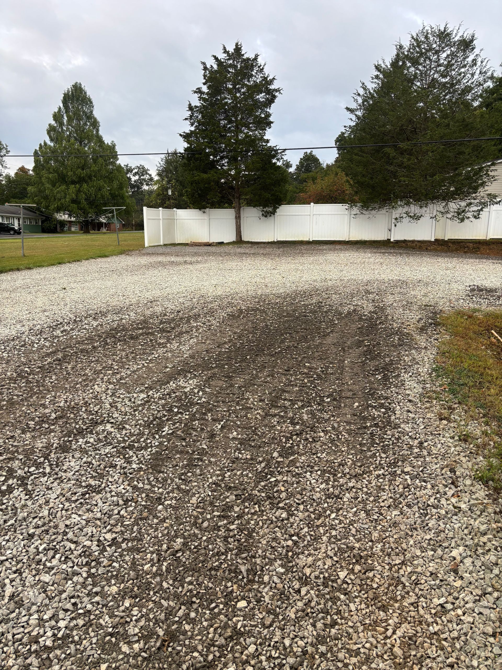 Gravel driveway leading to a white fence. Trees and overcast sky in the background.
