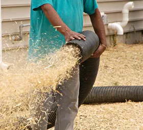 Man holding a black corrugated hose, expelling wood shavings outside.