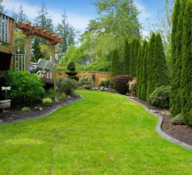 Green lawn with curved border, lush landscaping, wooden deck with pergola, tall evergreen trees.