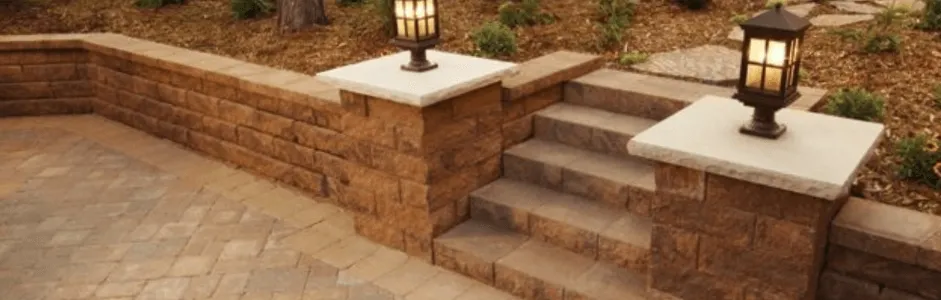 Brick patio with steps, retaining wall, and lanterns. Brown tones with plants in the background.