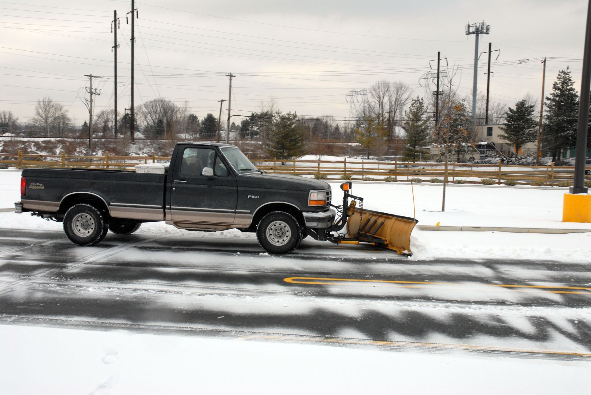 Dark pickup truck with a snowplow clearing a snow-covered parking lot on an overcast day.