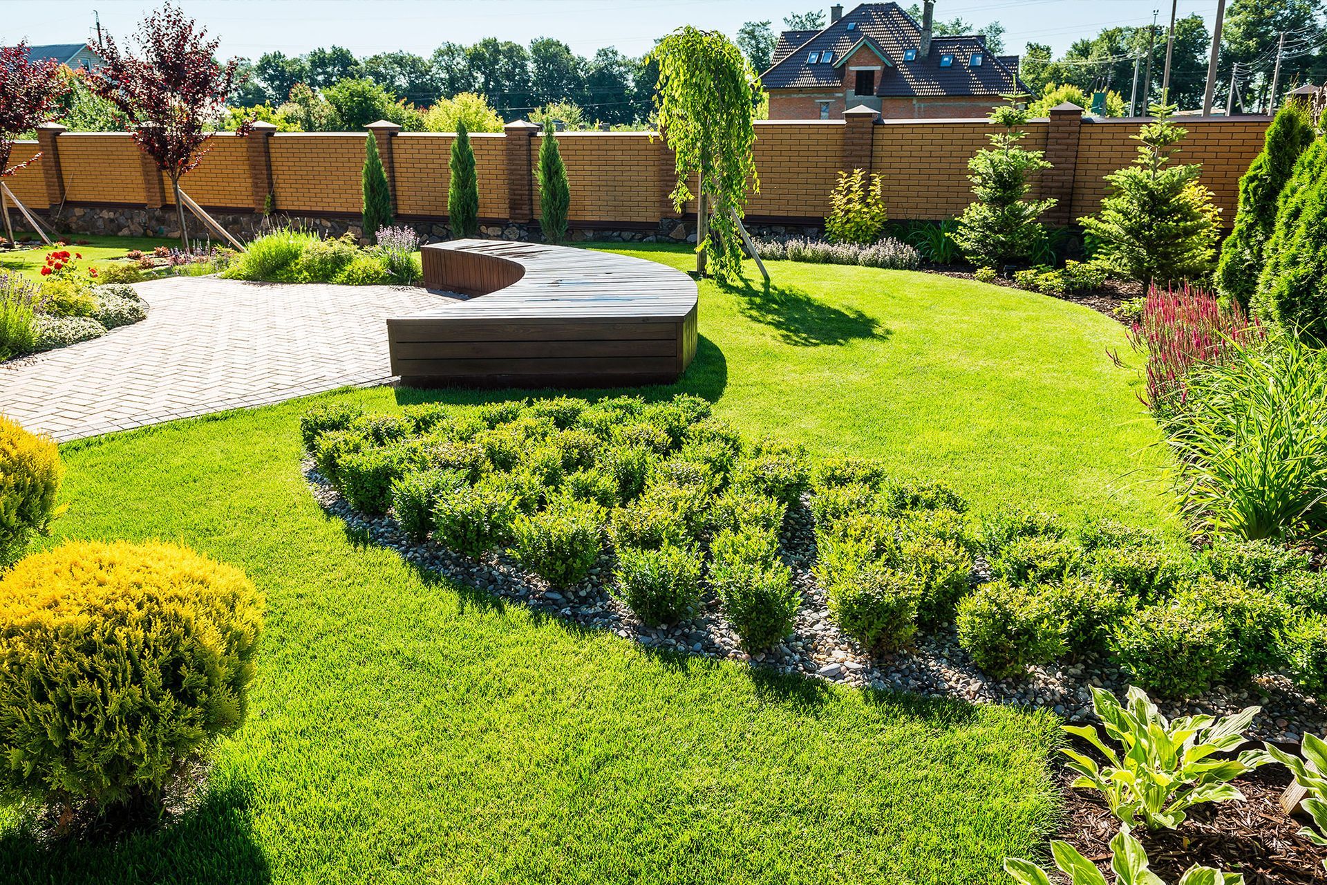 Lush green garden with manicured lawn, stone pathway, wooden bench, and various shrubs and trees under a sunny sky.