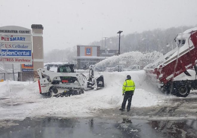 A skid-steer loader clears snow into a dump truck in a snowy shopping center parking lot near storefront signs.