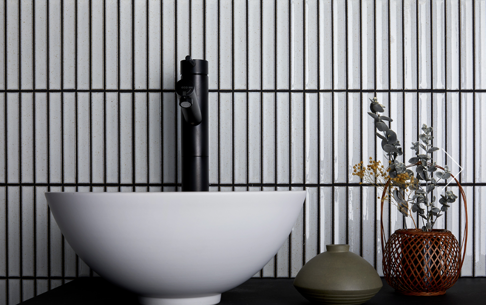 White bowl sink with black faucet against white and black vertical tiled wall, with decor.