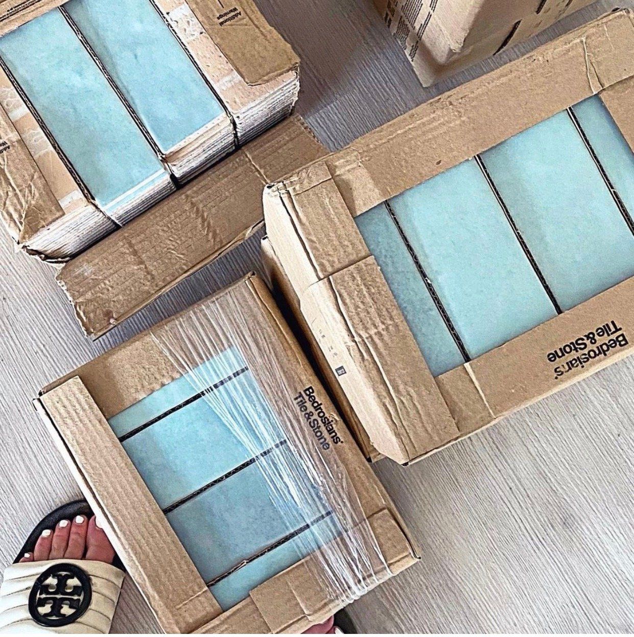 Boxes of light blue tiles, wrapped in plastic, on a light-colored floor. A foot wearing a white sandal is visible.