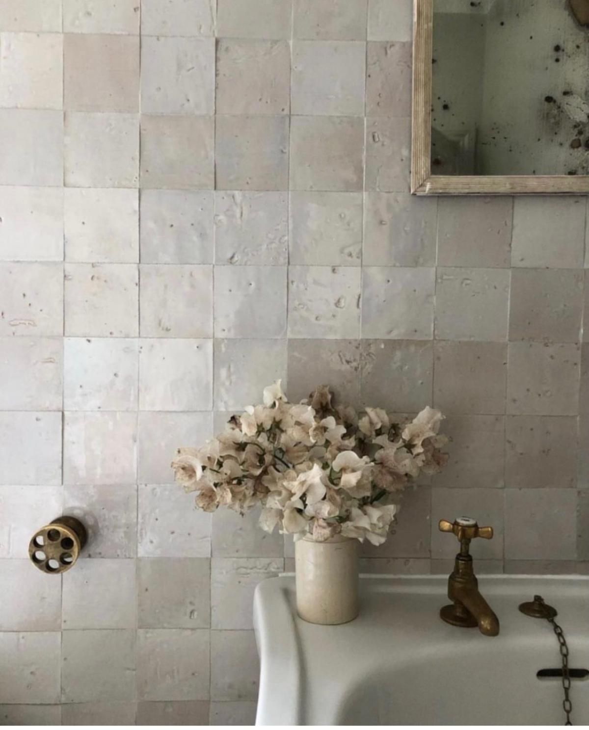 White tiled wall with a vase of flowers, brass faucet, and a vintage mirror.