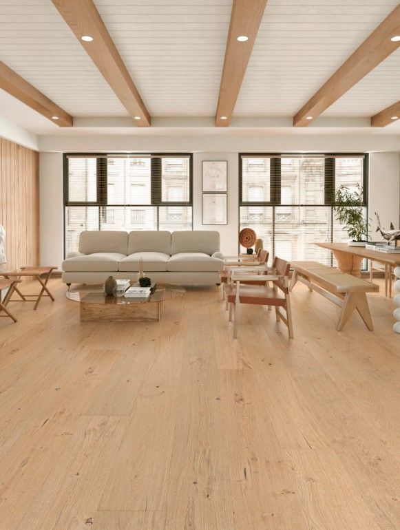 Modern living room with light wood floors, beige couch, large windows, and wooden beams.