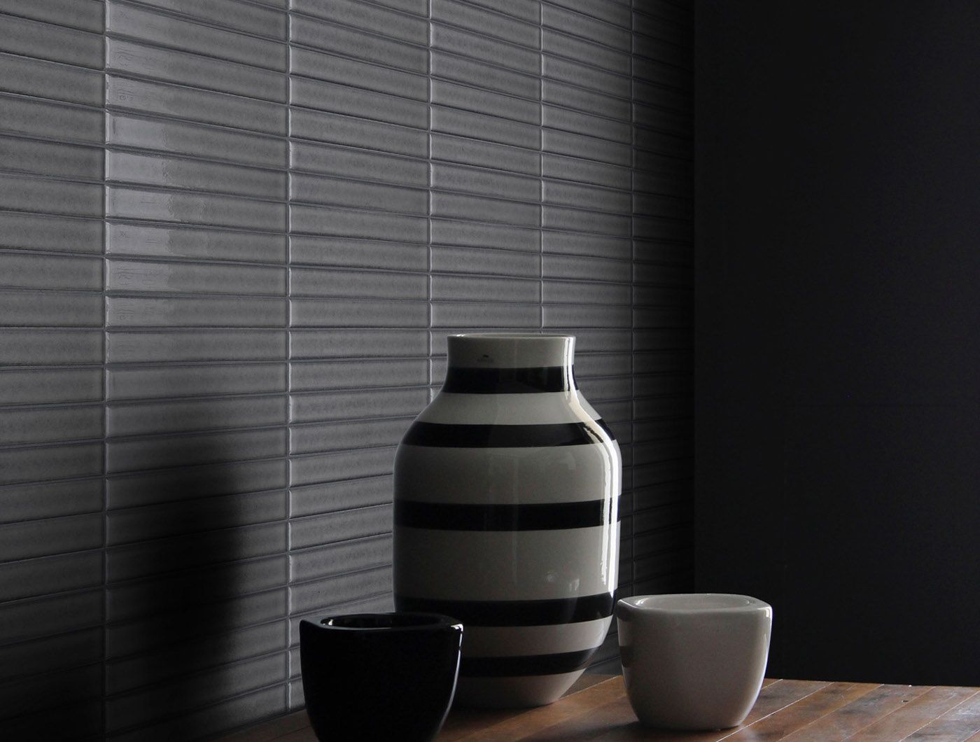 Dark grey tiled wall with striped vase and two cups on wooden table.