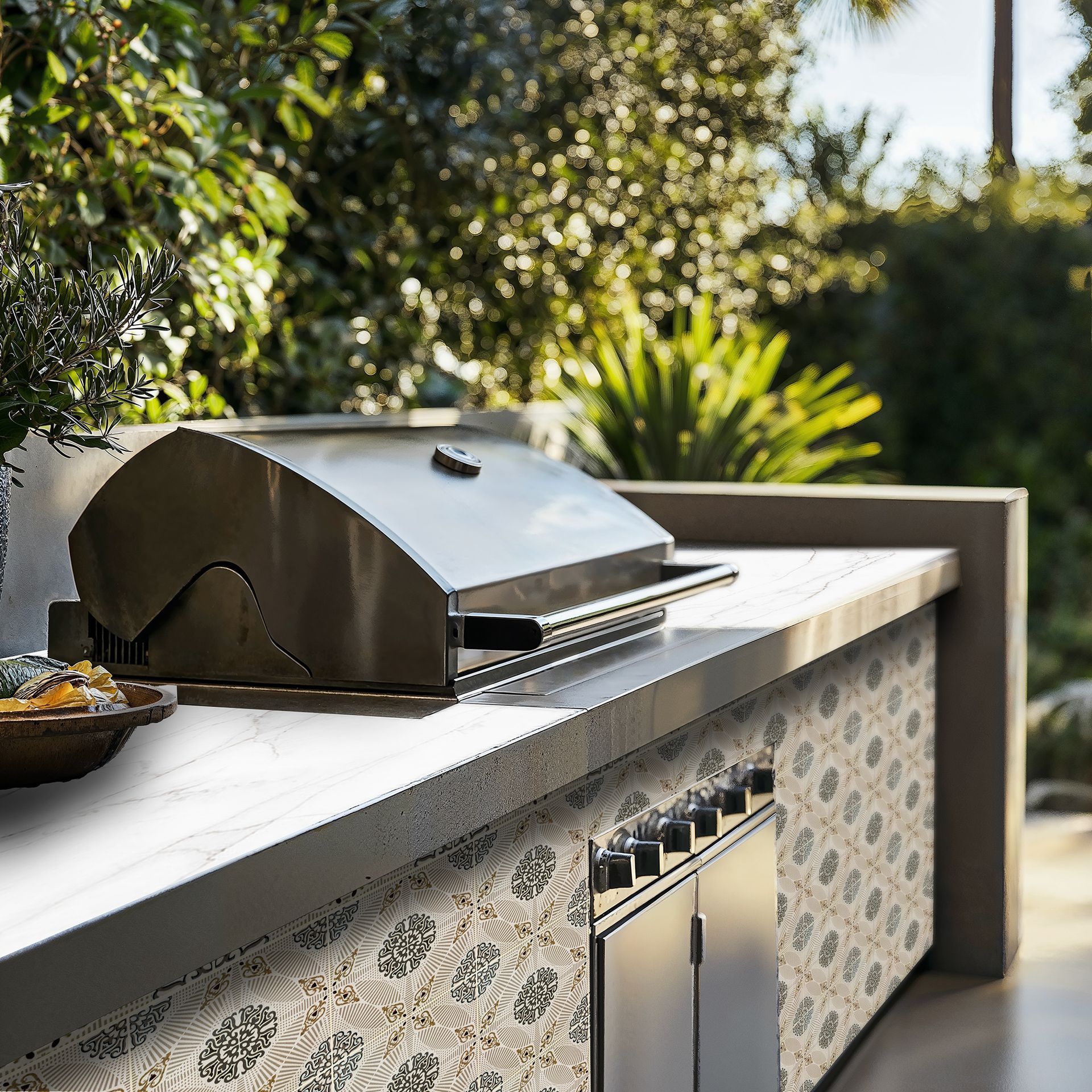 Outdoor kitchen with grill, tiled wall, and greenery in the background.