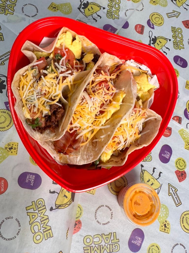 Three tacos in a red tray with cheese on a cartoon tablecloth, plus a small cup of orange sauce.