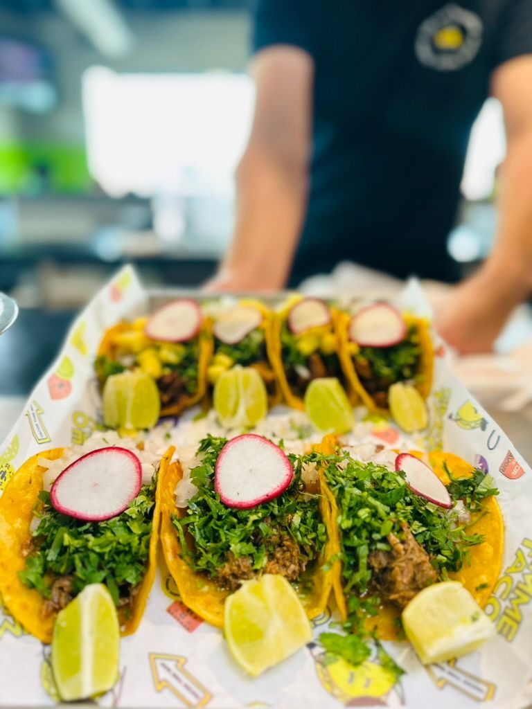 Tray of colorful tacos with radish slices and lime wedges, held by a person in a dark shirt.