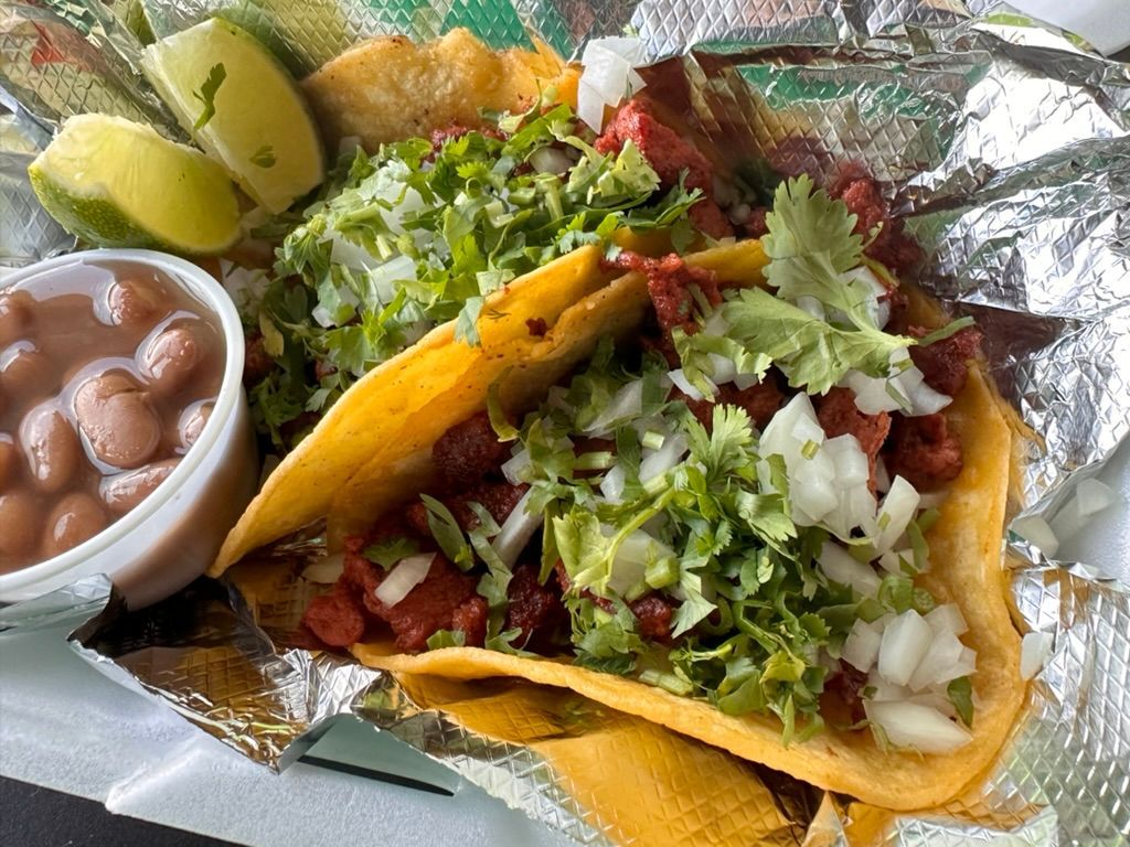 Two fish tacos with cilantro, onions, lime wedges, and a side of refried beans on foil wrapping
