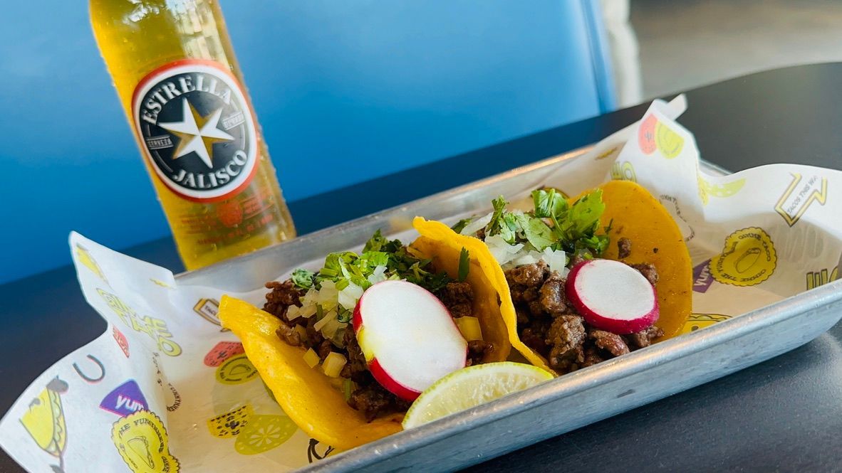 Two tacos in a paper tray with radish slices and cilantro, beside a beer bottle on a table.