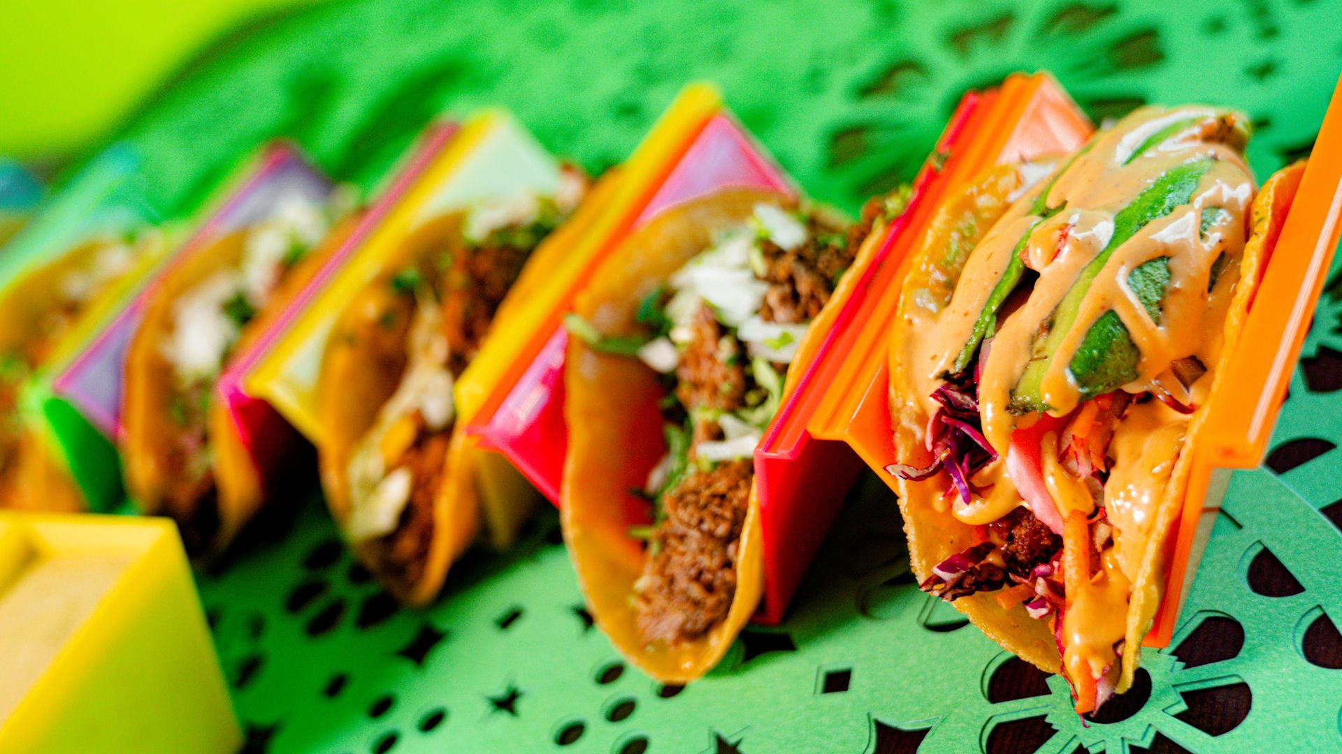 Colorful tacos lined up on a bright green perforated tray, filled with meat, vegetables, and sauce