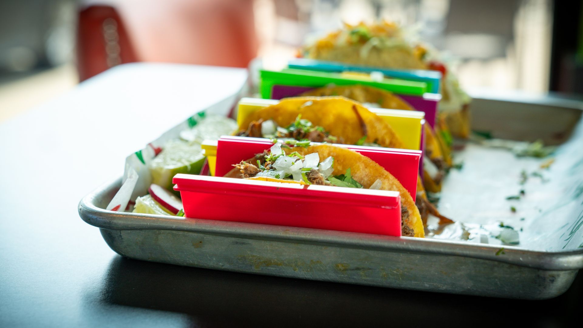 Tray of colorful tacos on a table, with red, yellow, and green shells topped with shredded garnish
