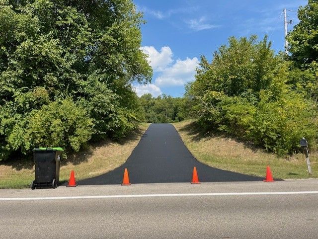 A black trash can sits on the side of a newly sealcoated road next to orange cones.