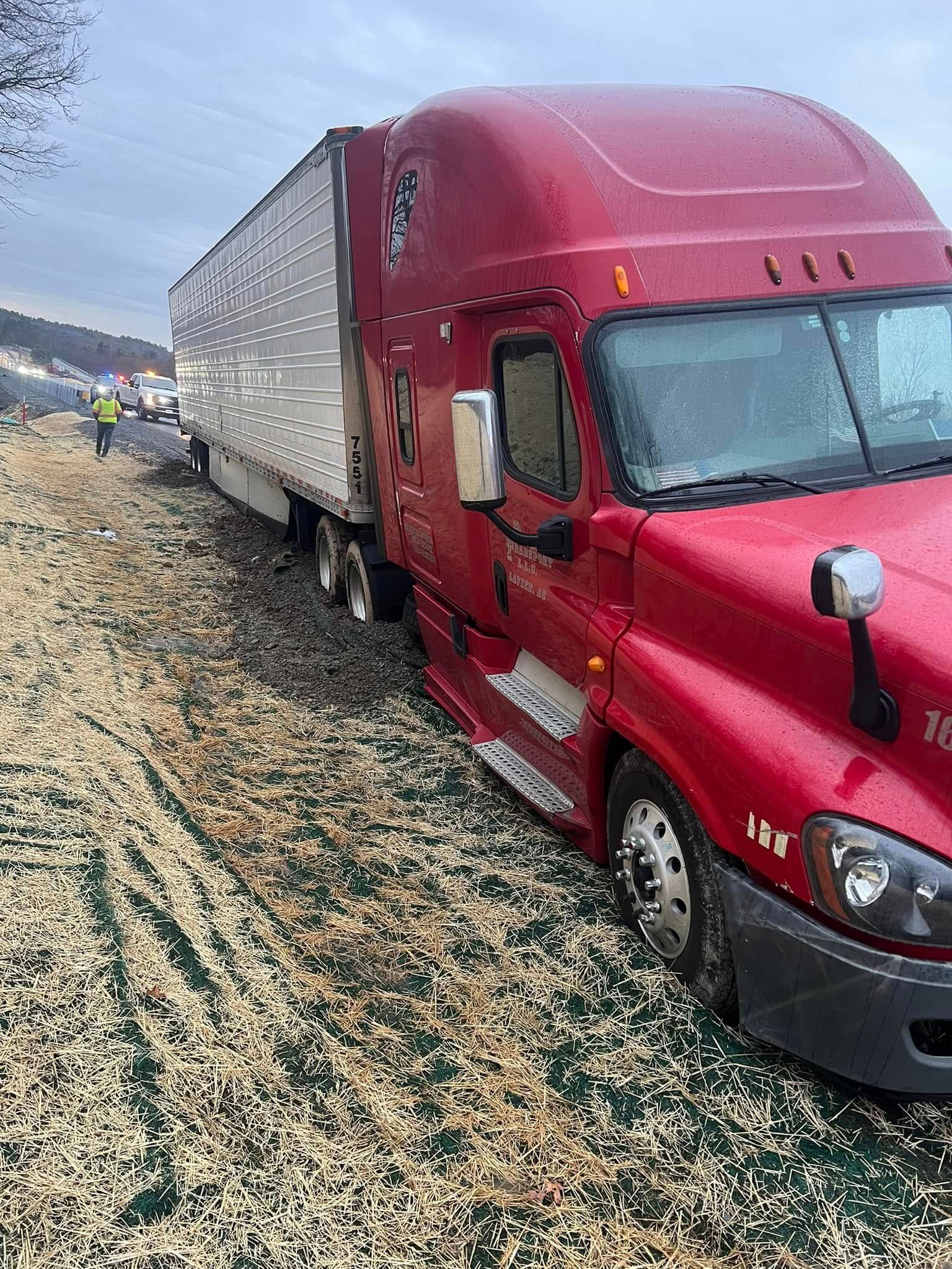 Red semi-truck off the road; white trailer, snow on the ground, emergency vehicle in the distance.