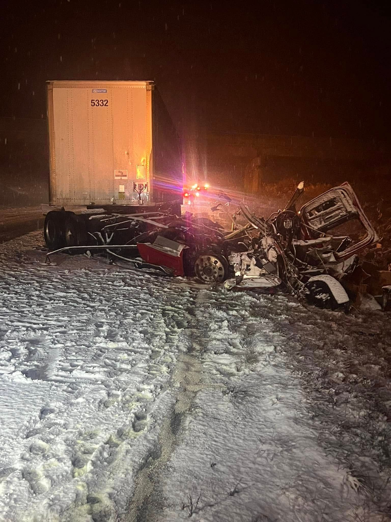 Semi-truck crashed on snow-covered road at night. The trailer is upright; the cab is wrecked. Emergency lights flash.