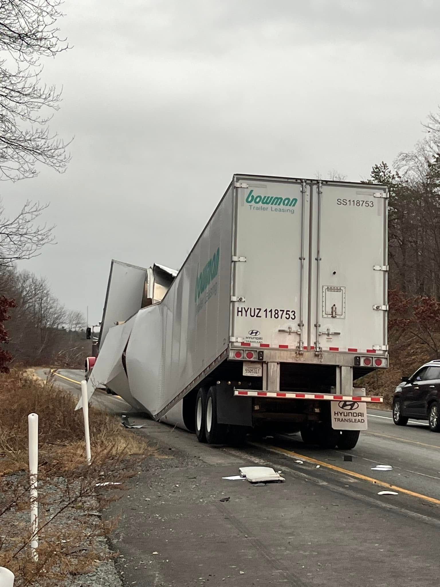 Semi-truck trailer severely damaged, crumpled on roadside. Gray sky, scattered debris.