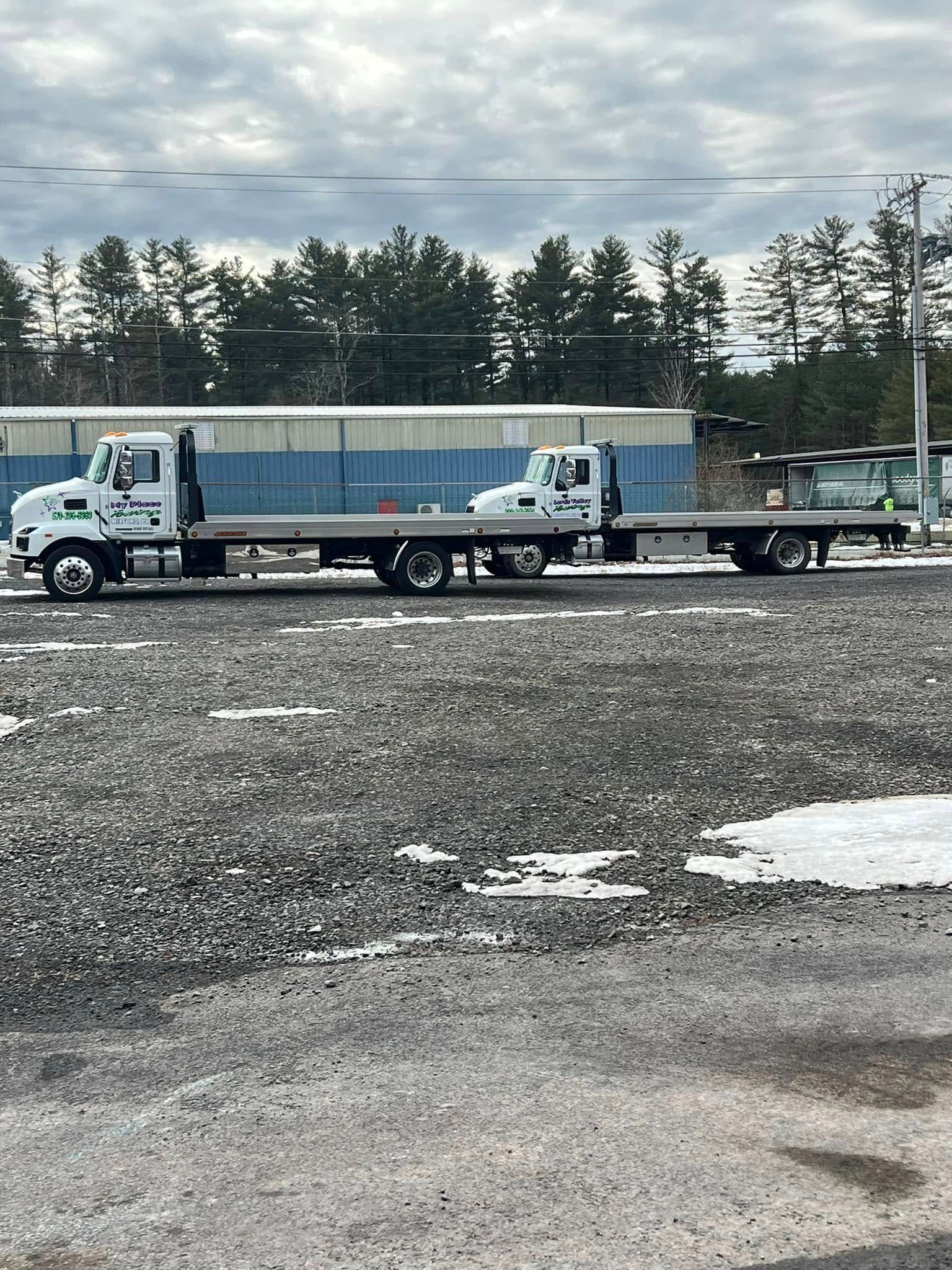 Two white flatbed tow trucks parked on gravel, with a backdrop of trees and a cloudy sky.