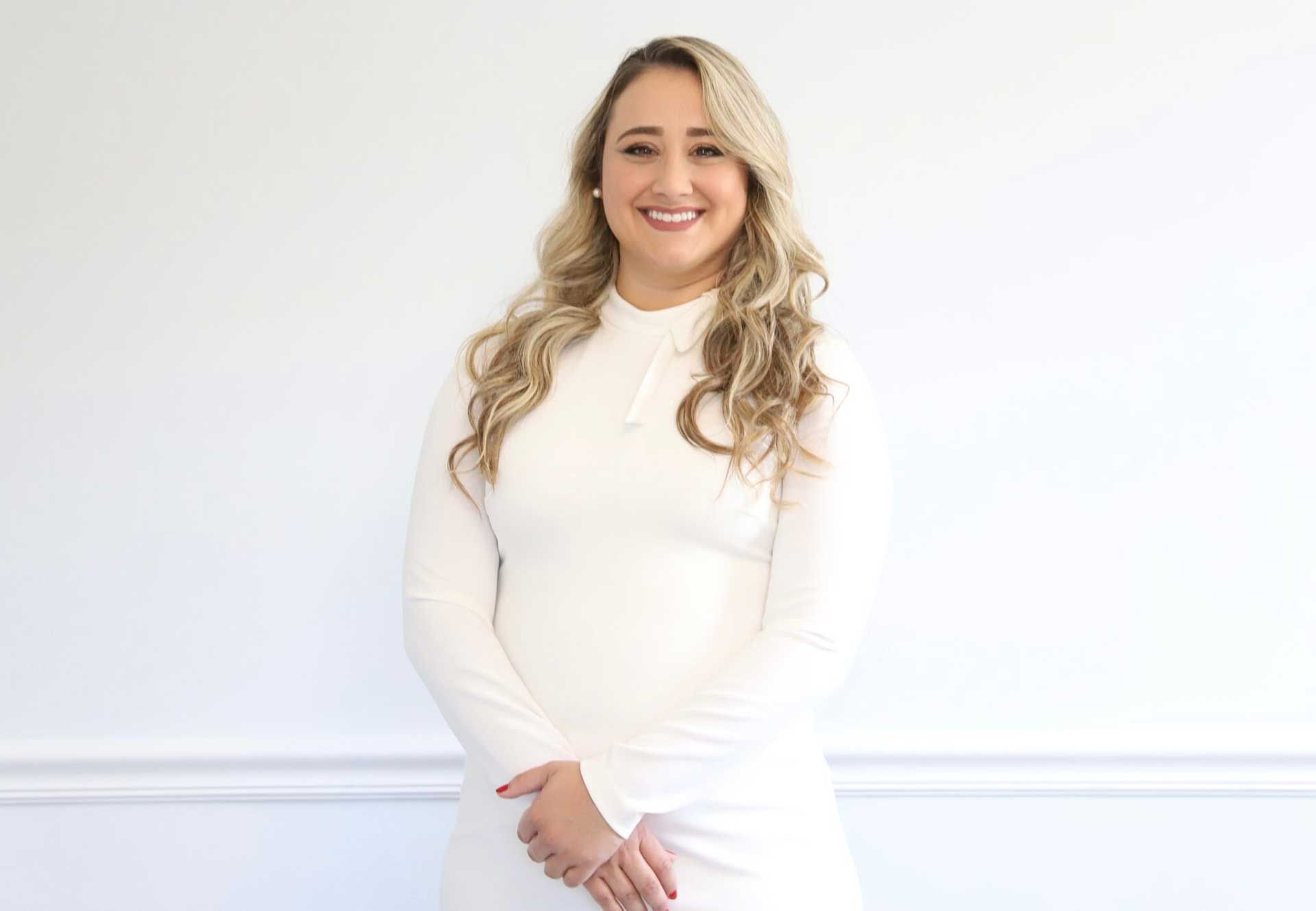 Blonde woman smiling, wearing a white dress, standing in front of a white wall.