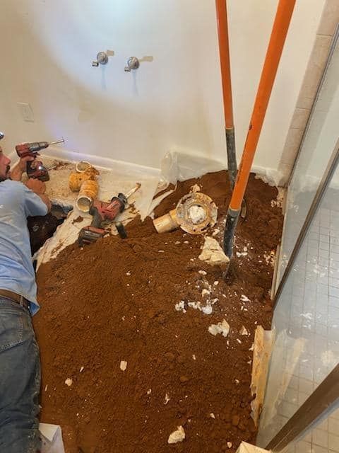 A man is working on a bathroom floor with a drill and shovel.
