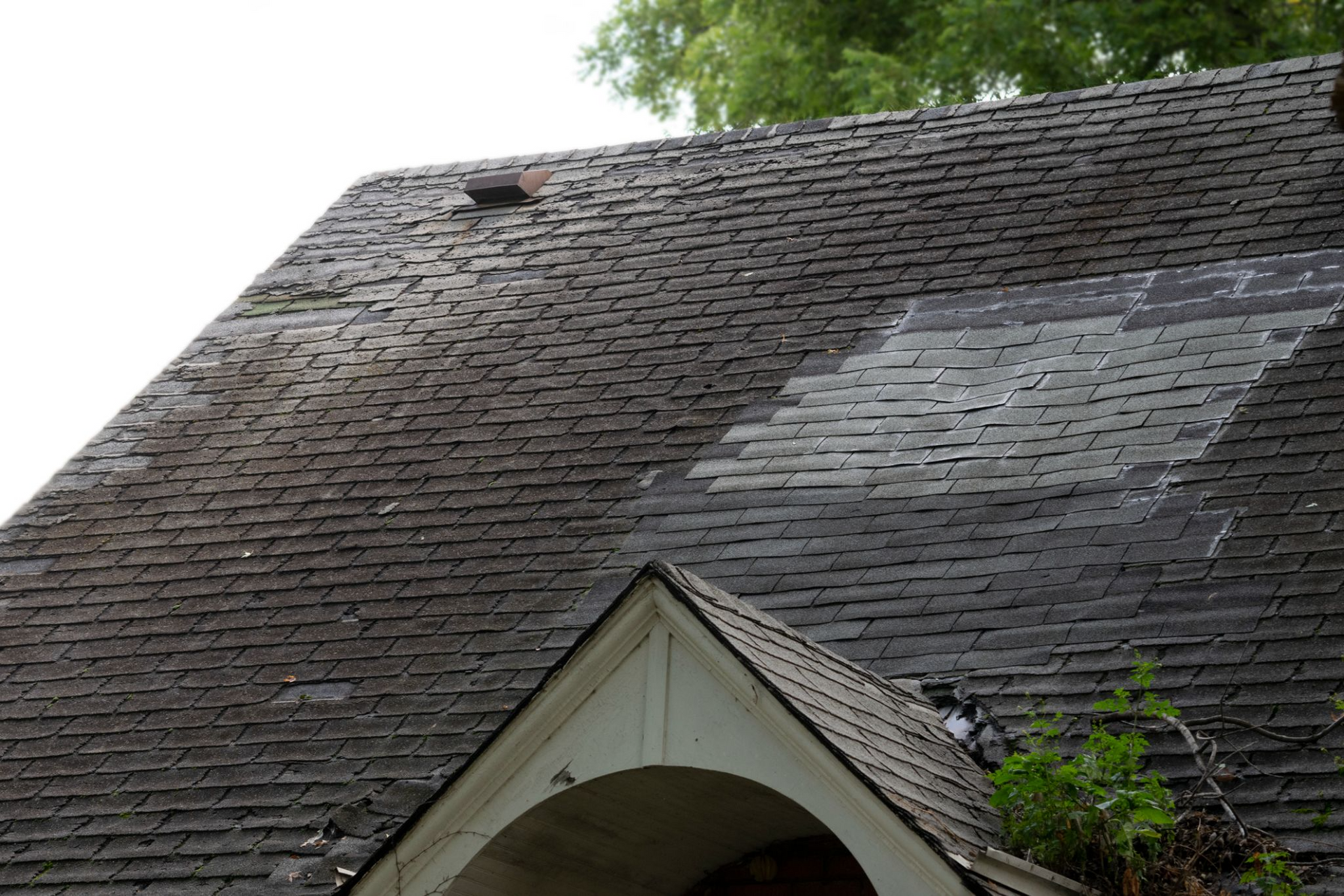 Damaged asphalt shingle roof, with missing and worn sections. A white gable with greenery below.