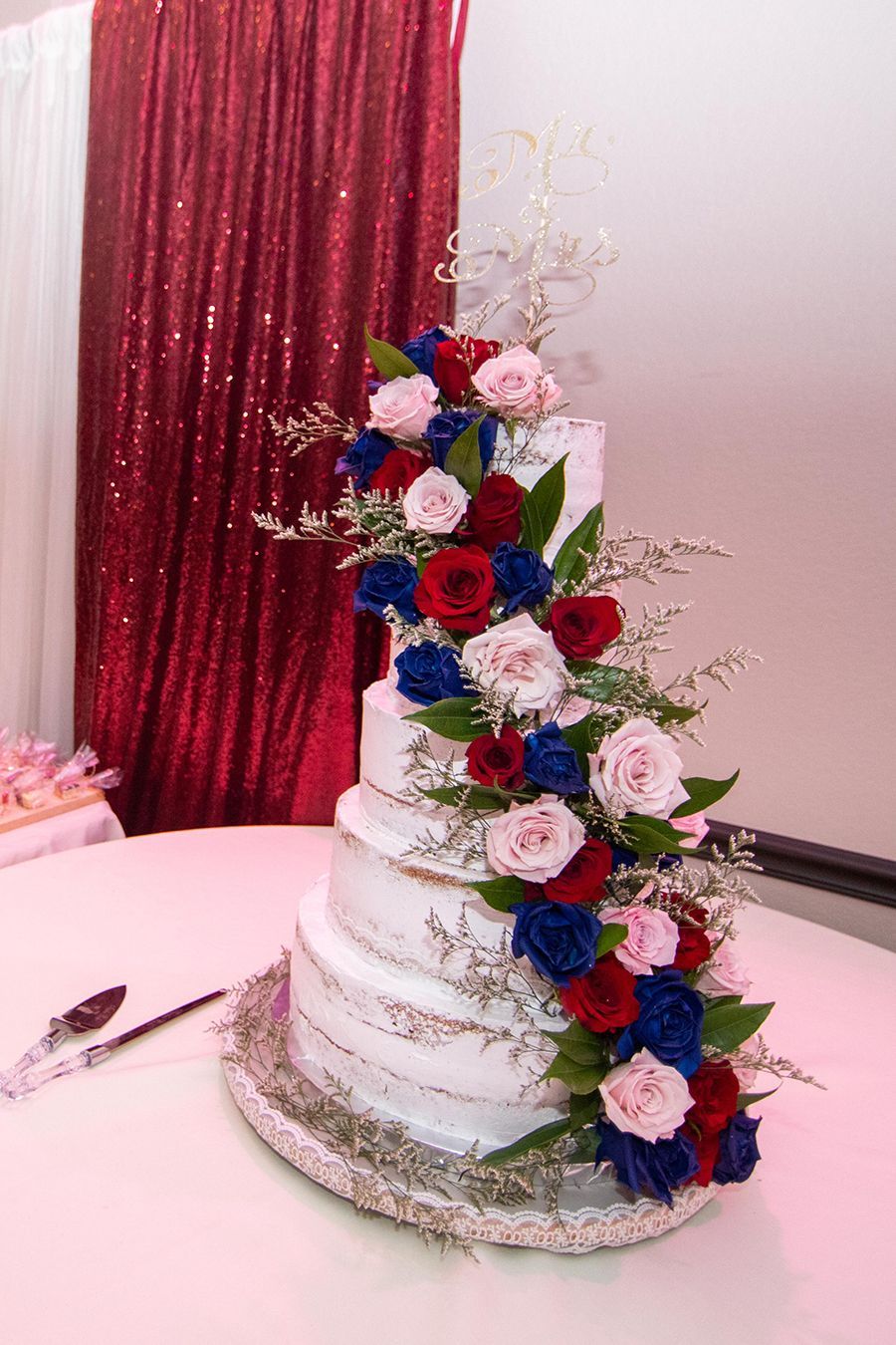 Five-tiered wedding cake decorated with red, blue, and pink flowers on a table with a red sequin backdrop.