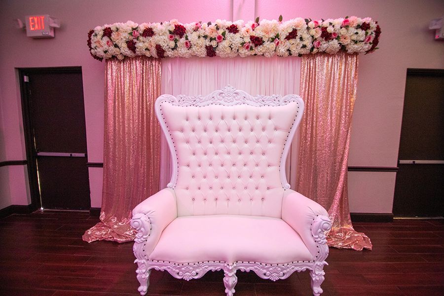 White ornate throne chair in front of a sequined backdrop with a floral arrangement.