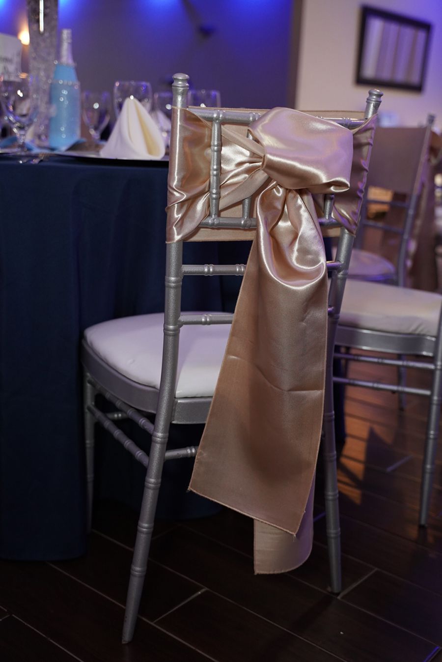 Silver chair with a champagne-colored satin bow tied to the back. A table setting is in the background.