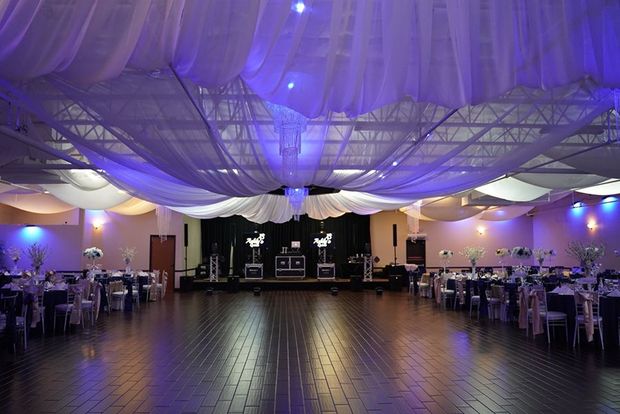 Reception hall with white draping ceiling, blue and white lighting, tables, and a stage.