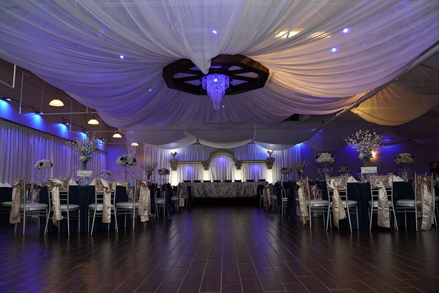 Elegant ballroom with draped ceiling, tables, chairs, and stage, bathed in blue and gold light.