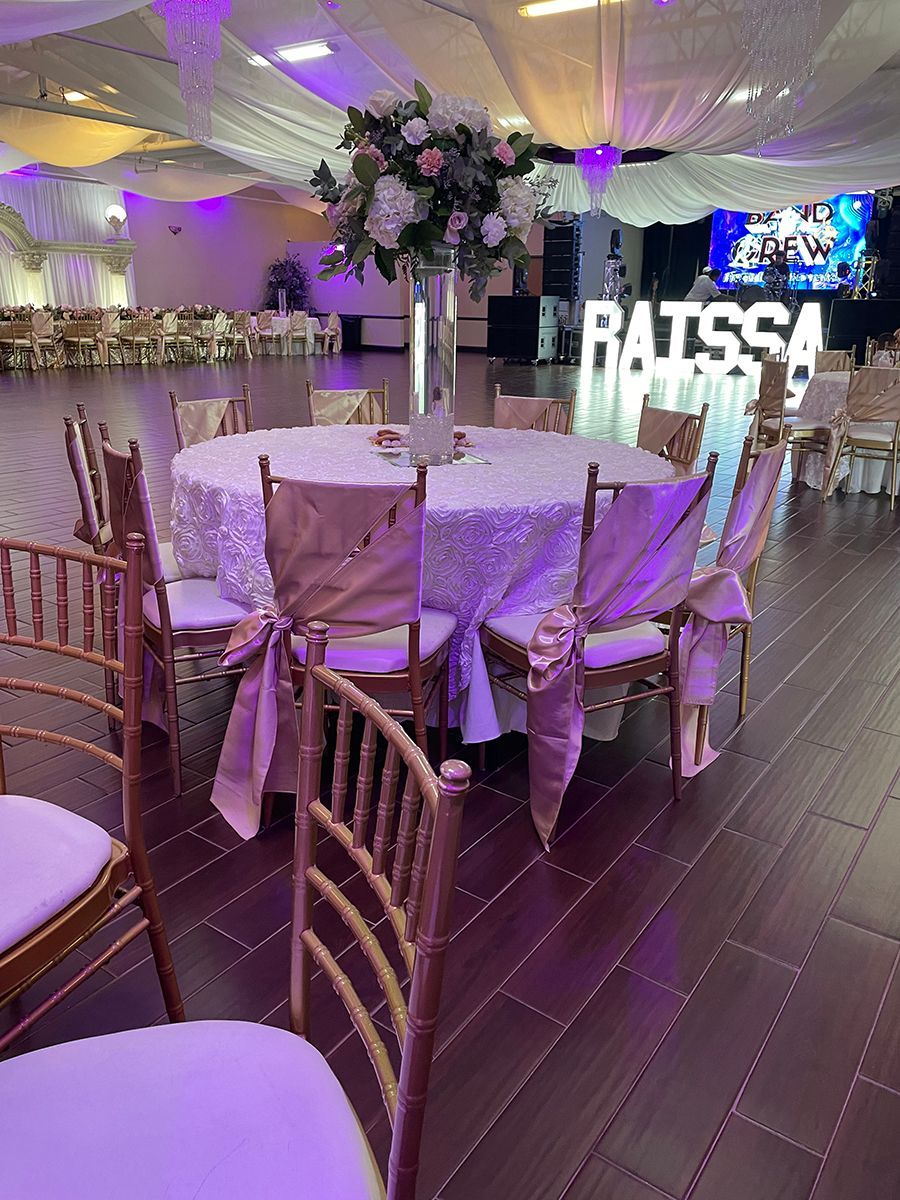 Round table set for a formal event with floral centerpiece and chairs decorated with ribbons.