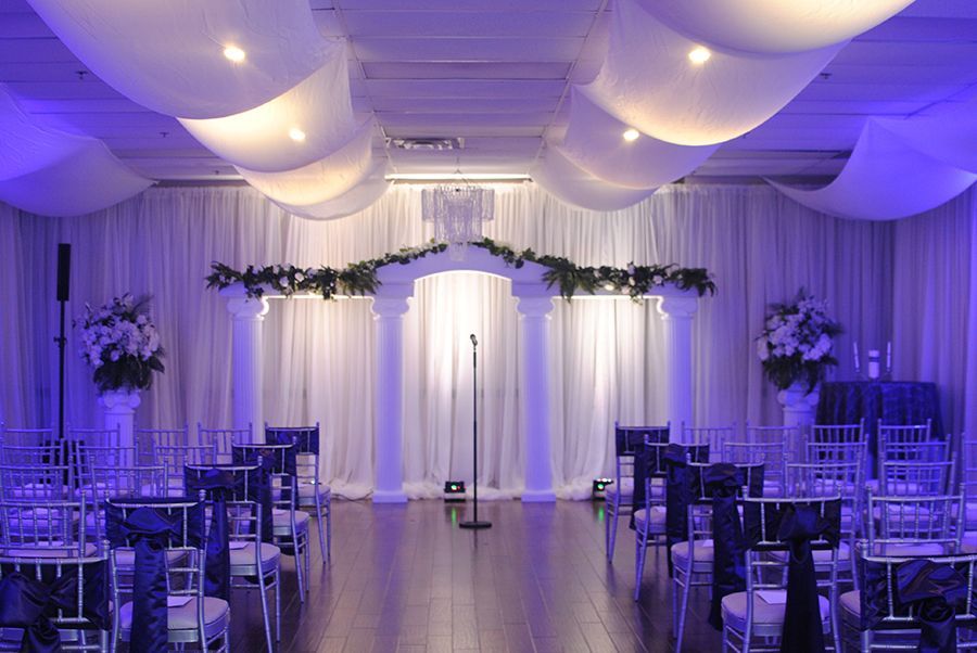 Wedding ceremony setup: white draped ceiling, altar with columns, blue chair sashes, and flower arrangements.