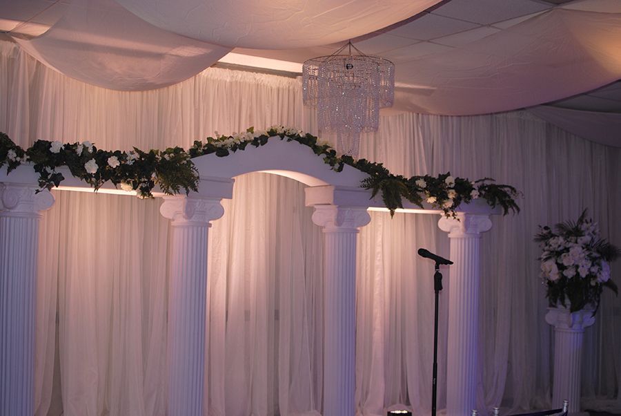 White wedding altar with draped fabric backdrop, floral garland, and chandelier.