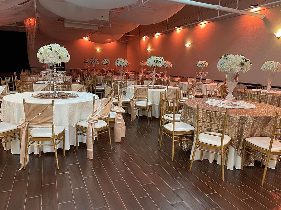 Elegant ballroom set for an event; round tables with gold decor and floral centerpieces; dark wood floor.