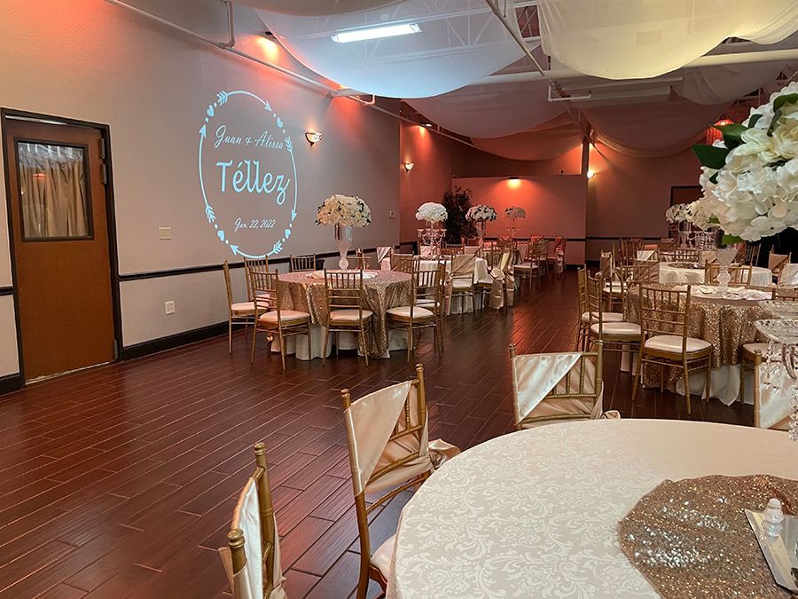 Wedding reception hall with round tables, gold chairs, and floral centerpieces. Name and date projected on the wall.